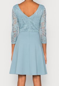 Robe bleu clair avec un corsage en dentelle et des manches trois-quarts. Jupe évasée et décolleté en V au dos. Texture lisse dans l'ensemble.