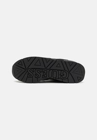Sort sneaker sål med en tekstureret gummioverflade, der har et trekantet mønster og "GUESS" logoet vist tydeligt.
