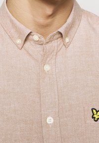 Lichtbruine overhem shirt van gestructureerde stof, met een kraag, parelachtige knopen en een geel geborduurd vlinderdetail.