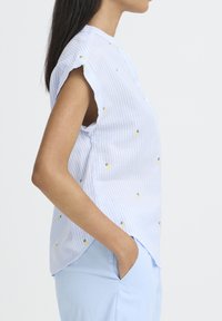 Femme en blouse sans manches rayée bleu et blanc avec de petits motifs ananas jaunes, portant un pantalon bleu clair, les mains dans les poches.