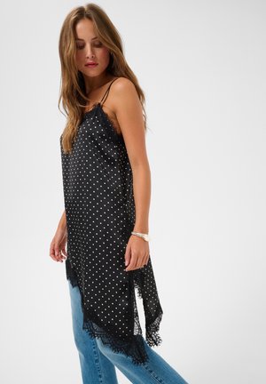 Femme portant une tunique noire sans manches à pois avec bordure en dentelle et un jean bleu clair, debout devant un fond clair uni.