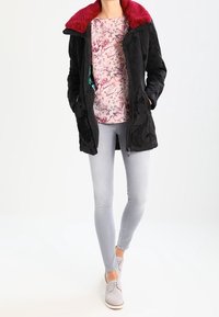 Veste noire avec un col en fourrure rouge, haut à motifs floraux, jean gris clair et chaussures grises. Présente des zips et des détails brodés.