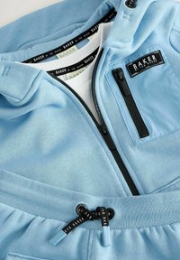 Svetle modrá mikina a kalhoty vyrobené z měkkého materiálu, s kapsou na zip, nastavitelnou šňůrkou a značkovými štítky v kontrastních barvách.