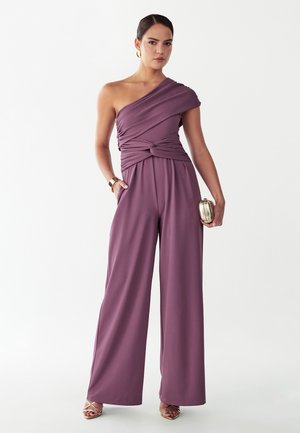 Žena v mauve overal s jedným ramenným remienkom a širokými nohavicami, drží striebornú kabelku, nosí zlaté náušnice, náramok a sandále.