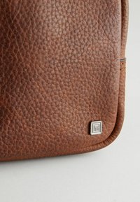 Angolo di una borsa in pelle marrone con textura, impreziosito da un accento in logo argentato. Superficie morbida e gommosa con bordi arrotondati, caratterizzata da dettagli di cucitura.