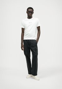 Balta kokvilnas t-krekls ar maziem melniem tekstiem; kombinēts ar melnām kravas biksēm ar sānu kabatām; stilizēts ar bēšiem slip-on apaviem.