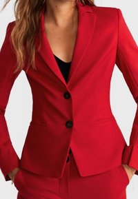 Kvinde iført en tætsiddende rød blazer med sorte knapper over en sort top, med hænderne i matchende røde bukselommer.
