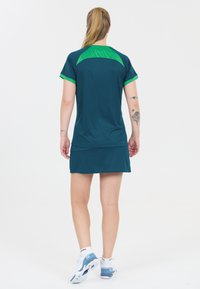 Teal sportskjole med grønne accenter. Korte ærmer og en tætsiddende design. Båret med hvide og blå atletiske sko og synlige tatoveringer på underarmen.