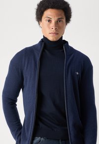 Marinblå zip-up cardigan med hög krage och ribbade ärmslut, lager på lager över en mörk turtleneck sweater, tillverkad av en mjuk tygblandning.