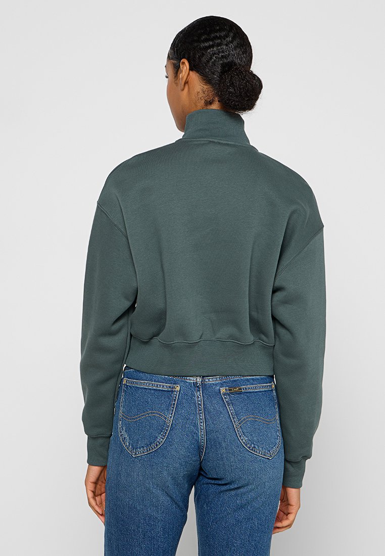 Sudadera corta de color verde oscuro con cuello alto y mangas raglán, que presenta una textura suave y un dobladillo acanalado, combinada con jeans azules.