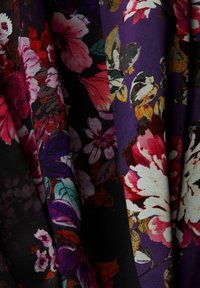 Tissu avec des motifs floraux denses dans des tons rose, rouge, blanc et violet sur un fond sombre, montrant des plis et de la texture.