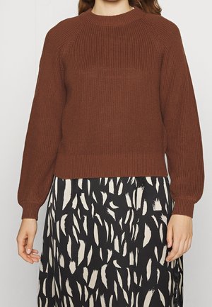 Kobieta ubrana w brązowy sweter z dzianiny i czarną spódnicę z białymi abstrakcyjnymi wzorami pędzla, stojąca na tle jednolitego tła.