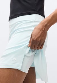 Ανοιχτά μπλε αθλητικά σορτς με πλευρικές σχισμές, κατασκευασμένα από μαλακό ύφασμα. Ένα λευκό λογότυπο φαίνεται στο κάτω αριστερό μέρος. Χέρι κρατά το τελείωμα.