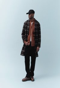 Čovjek u crnoj kapi, smeđem džemperu na patentni zatvarač, karo kaputu, crnim hlačama i smeđim cipelama stoji ispred svijetloplave pozadine.