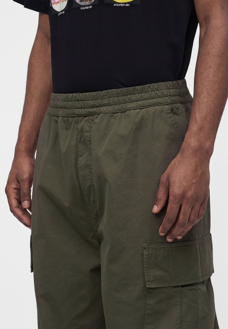 Mann trägt olivgrüne Cargo-Shorts mit elastischem Bund und Seitentaschen, kombiniert mit einem schwarzen T-Shirt mit Grafik.