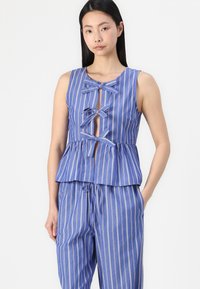 Modra in bela črtasta peplum majica z ključavnico spredaj, ki ima tri mašne zavezovanje in zaobljen izrez. Tkanina ima gladko teksturo.