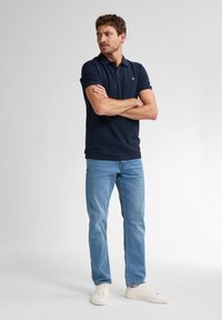 Marineblauwe poloshirt met korte mouwen, gecombineerd met lichtblauwe jeans en witte sneakers, staand op een effen achtergrond.