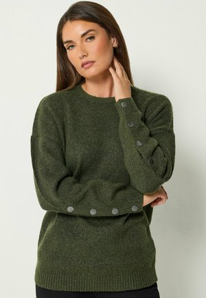 Mujer con cabello castaño liso que lleva un suéter verde oscuro con cinco botones plateados en cada manga sobre un fondo claro.