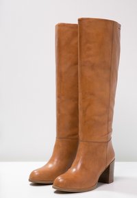 Bottes en cuir marron jusqu'au genou avec une texture lisse, un bout rond et un talon épais, présentant des coutures minimales et une finition polie.