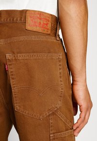 Pantalon en denim marron avec un patch en cuir arborant un logo rouge. Les poches arrière sont ornées d'une couture courbée. Texture lisse et matériau robuste.