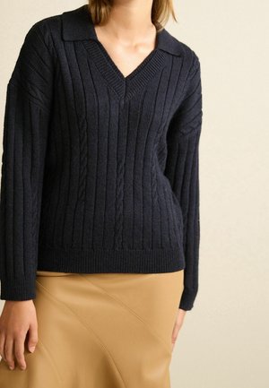 Femme portant un pull en maille torsadée bleu marine foncé à col en V, associé à une jupe en cuir beige, debout devant un fond neutre.