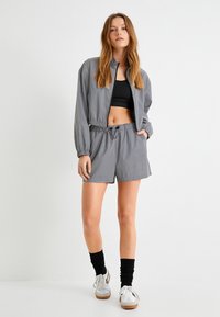 Szara sportowa kurtka z zamkiem błyskawicznym z przodu, krótkie spodenki z elastycznym pasem, czarny top w stylu crop oraz czarne skarpetki do kostek, zestawione z białymi sneakersami.