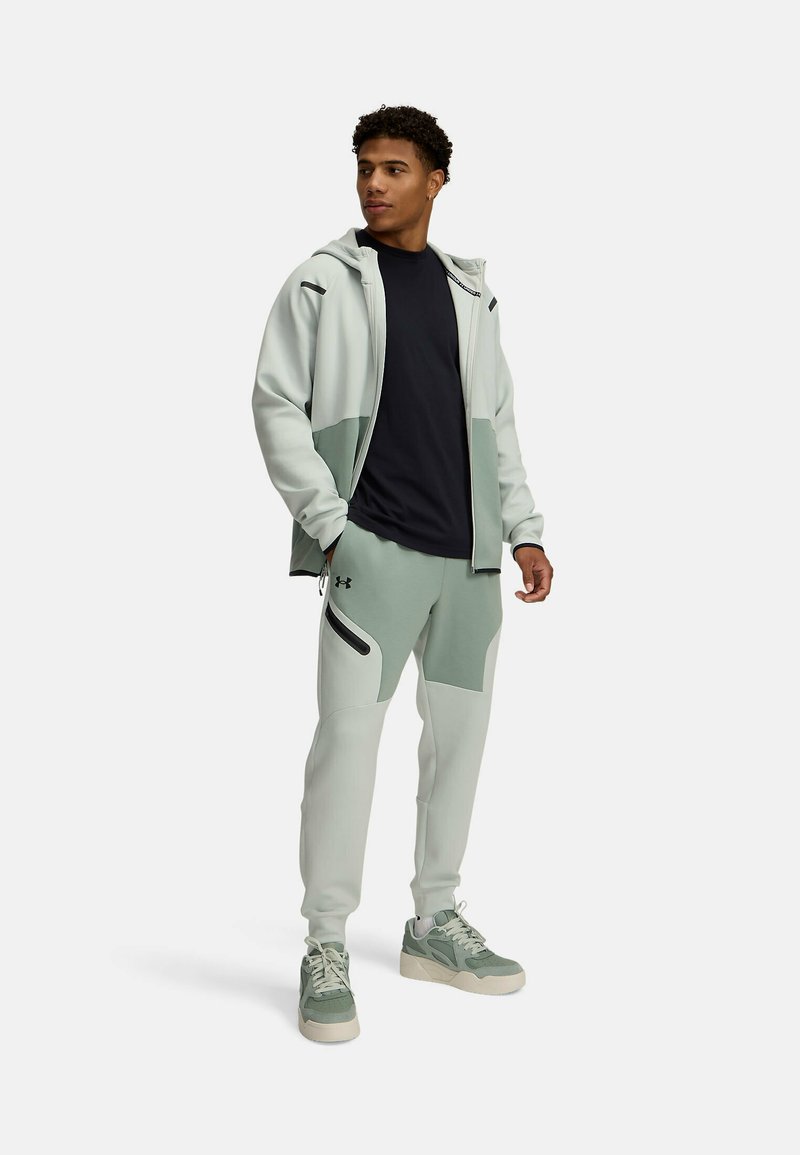 Haut de sport gris clair avec des accents vert foncé, associé à un jogging assorti avec des poches latérales. Porté avec des baskets vertes.