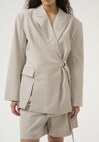 Heller beiger Wickelblazer aus strukturiertem Stoff mit einem subtilen Karomuster, langen Ärmeln, einem Gürtel und einem passenden Rock.