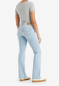 Personne portant un jean droit bleu clair, un haut gris à manches courtes et des baskets blanches, debout avec le dos légèrement tourné.