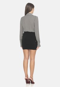 Mini gonna nera con dettaglio di bottoni laterali, abbinata a una blouse a maniche lunghe stampata. La modella indossa scarpe col tacco bordeaux.