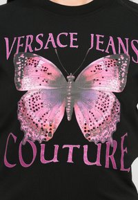 Svart t-skjorte med en stor sommerfugl grafikk i rosa og lilla toner med rhinstein detaljer. "Versace Jeans Couture" tekst over og under.
