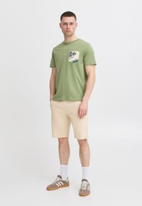 Zelené bavlněné tričko s vzorovanou kapsou, béžové šortky a hnědé tenisky. Stojící postava s krátkými vlasy a bílými ponožkami.