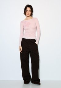 Frau mit kurzen dunklen Haaren, die ein hellrosa Langarmshirt mit floralen Details und lockere schwarze Hosen trägt, posiert vor einer schlichten weißen Wand.