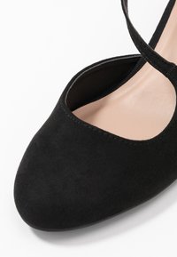 Primer plano de un zapato de tacón alto de ante negro con puntera redondeada y correa en el tobillo, sobre un fondo blanco.
