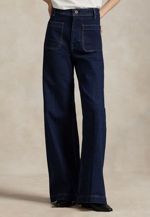 Blugi de culoare albastru închis, cu talie înaltă și picior evazat, buzunare mari aplicate în față și închidere cu un singur nasture, purtați cu pantofi negri cu vârf ascuțit.