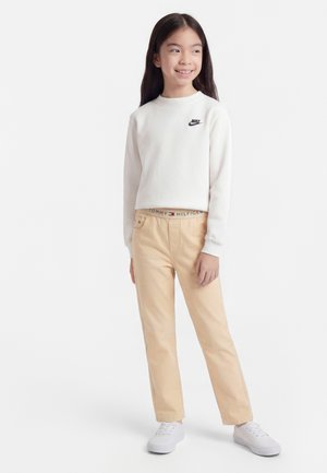 Κορίτσι με μακριά σκούρα μαλλιά φοράει λευκό φούτερ Nike, μπεζ παντελόνι Tommy Hilfiger και λευκά αθλητικά παπούτσια, στέκεται με χαλαρά χέρια.