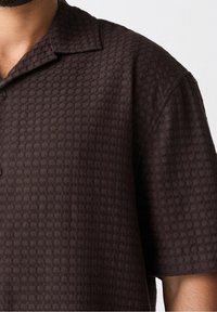 Camisa de manga corta en textura de gofre color marrón oscuro, con cuello pointed y cierre de un solo botón, de corte relajado y cuadrado.