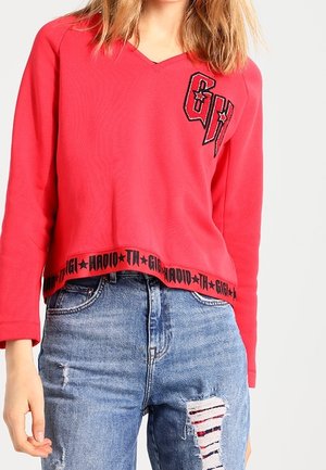 Sweat-shirt rouge court avec un col en V, orné d'un logo noir brodé et de texte au bas. Associé à un jeans bleu déchiré.