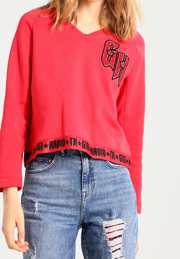 Rode cropped sweatshirt met V-hals, voorzien van een zwart geborduurd logo en tekst aan de zoom. Gepaarde met versleten blauwe jeans.