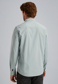 Camisa de manga larga azul claro con cuello abotonado, textura suave y dobladillo curvo; cuenta con puños abotonados y detalles de costura sutiles.