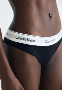 Tanga preta com uma cintura elástica branca que apresenta o logótipo "Calvin Klein". Textura suave com um design minimalista.