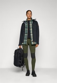 Zwarte waterdichte jas over een geruit groen en crèmekleurig shirt, zwart topje, olijfgroene leggings met kniestukken en houdt een zwarte rugzak vast.