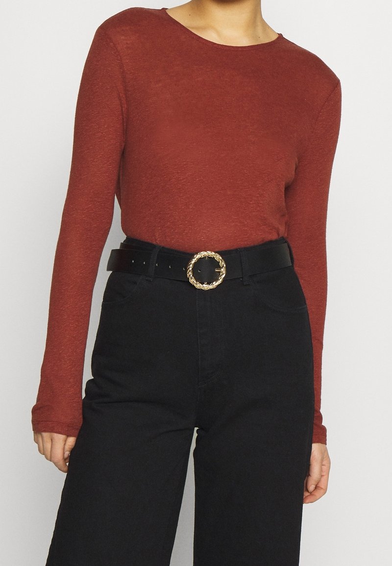 Långärmad rostfärgad topp kombinerad med högmidjade svarta jeans och ett svart bälte med en detaljerad, flätad guldspänne. Slät tygtextur.