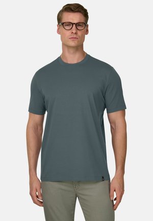 Mężczyzna o krótkich brązowych włosach i okularach, ubrany w gładki ciemnoturkusowy t-shirt z krótkim rękawem oraz jasne spodnie, stojący na jasnym tle.