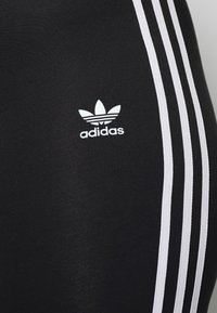 Leggings atletici neri con tre strisce verticali bianche lungo i lati. Presentano un piccolo logo Adidas bianco vicino all'area dell'anca.