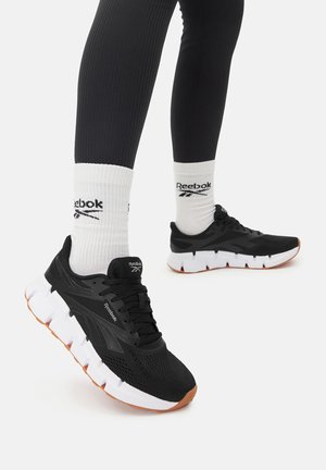 Zapatillas de running negras Reebok con suelas blancas gruesas, usadas con calcetines blancos Reebok de caña alta y leggings negros sobre un fondo blanco.