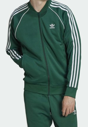 Persoon die een groen Adidas trainingspak draagt met witte strepen op de mouwen en broek, staand met één hand in de zak en de andere ontspannen.