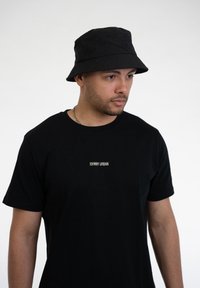 Svart bucket hat i tyg med en subtil logodetalj. Bärs med en svart t-shirt utan mönster som har liten vit text. Mjuk textur.