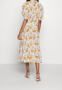 Femme portant une robe midi blanche à manches bouffantes et imprimé floral orange, associée à des chaussures slingback à talons bas beiges, se tenant de dos.