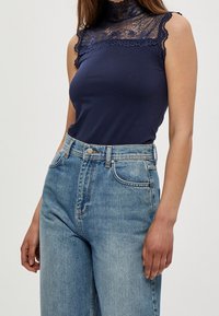 Haut sans manches bleu marine avec un col en dentelle haut, associé à un jean taille haute bleu clair avec une fermeture à boutons et à fermeture éclair classique.